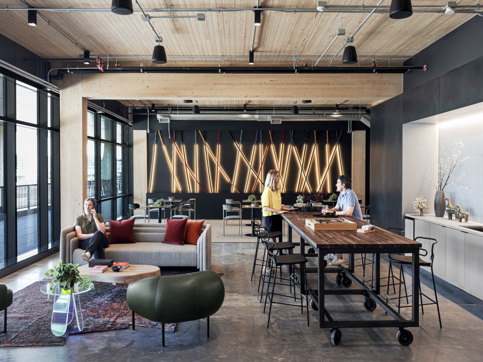 interior of a mass timber building; floor to ceiling windows with steel frame and mass timber elements exposed; gray concrete floor; people in a communal kitchen space with varied seating talking; Right wall with sink faces floor to ceiling windows with patio space; backwall lighting design elements feature long sticks of light laid on top of each other