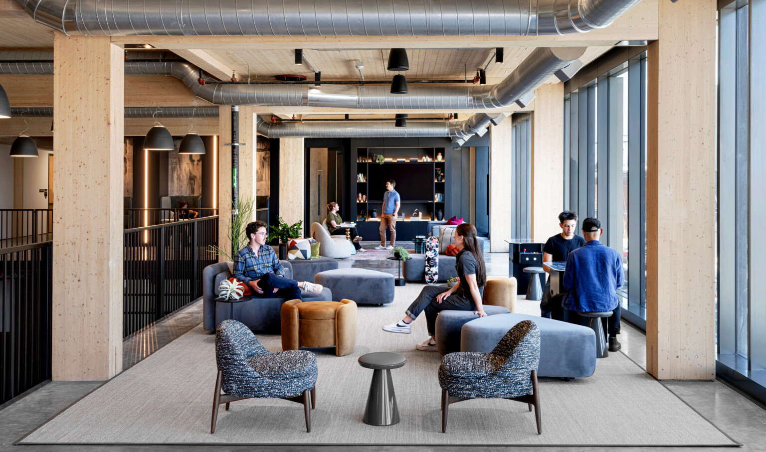interior of a mass timber building; floor to ceiling windows with steel frame and mass timber elements exposed; gray concrete floor; people in a communal space with varied seating talking; back wall entrances to workspace and bookshelf design elements