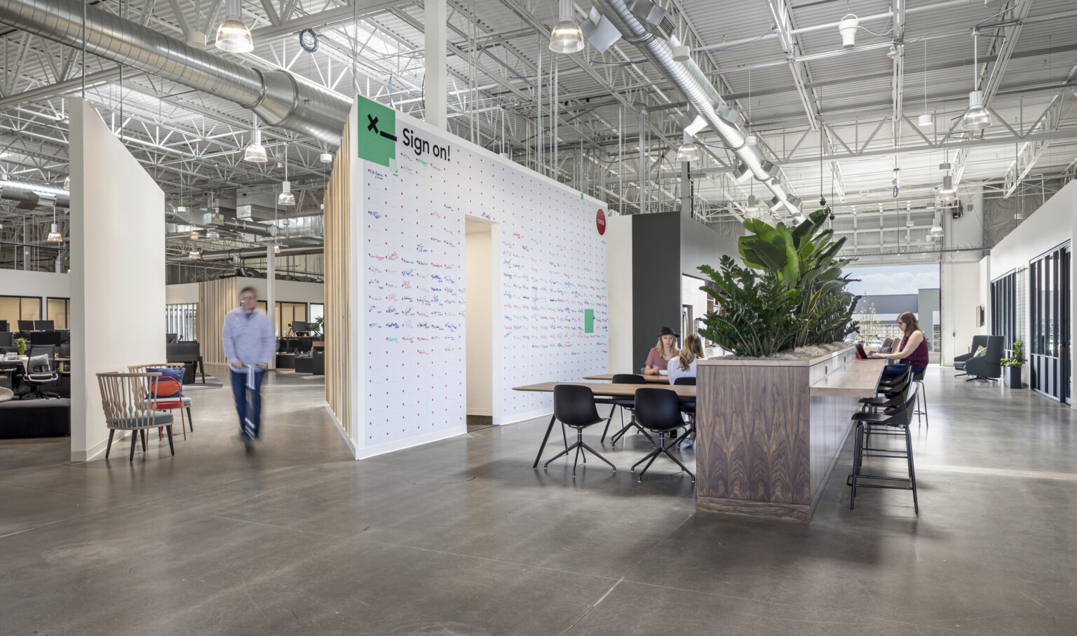 Tables in front of wood half wall with plants on top and counter attached. White board wall to left. Open garage door window