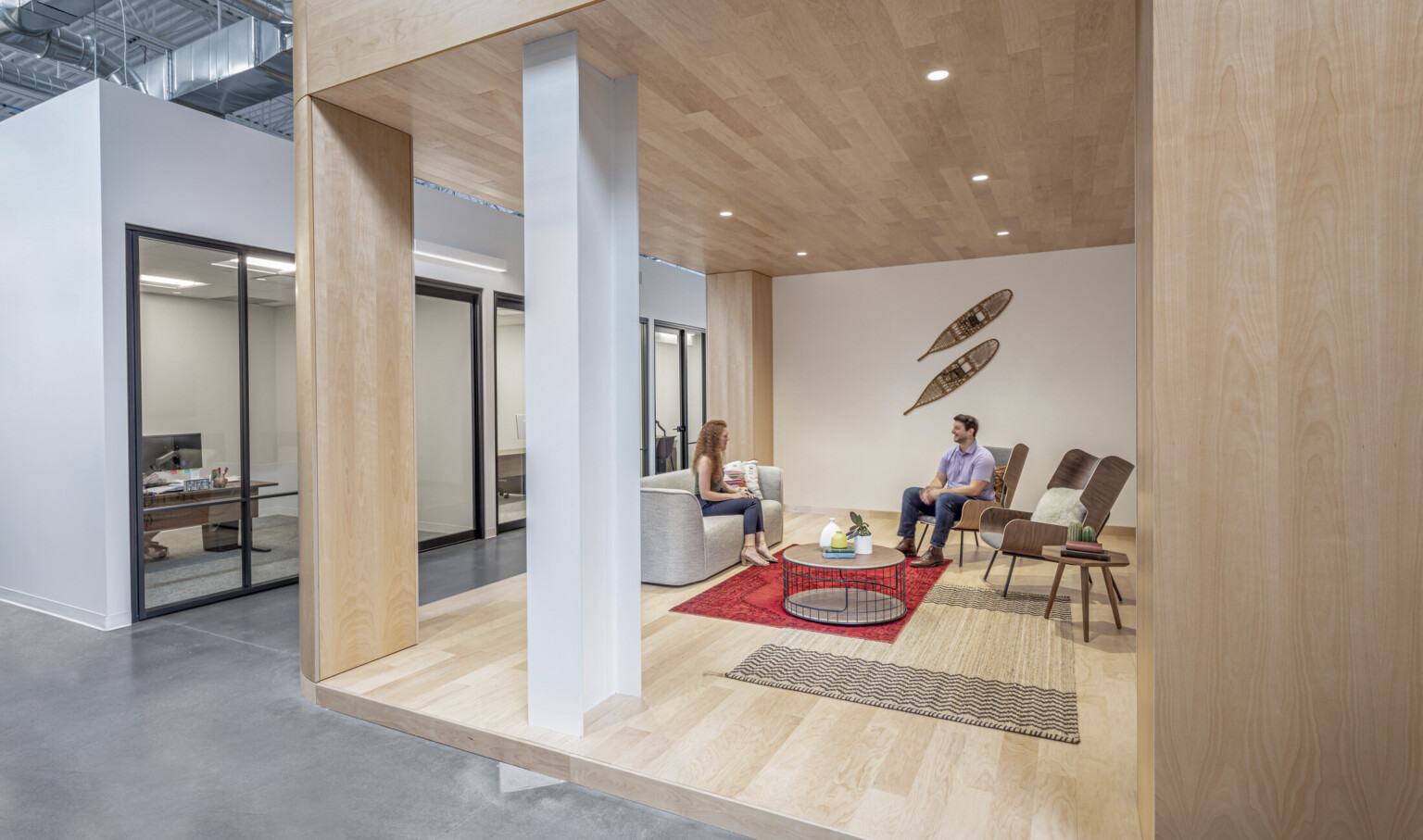 Wood alcove opposite glass walled offices in white hallway. Decorative snowshoes on wall above seating area framed by columns
