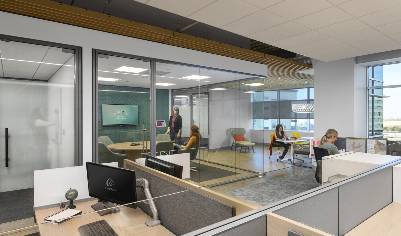 Cubicles with glass partitions in space lined with glass walled work rooms with seating, tables, and screens