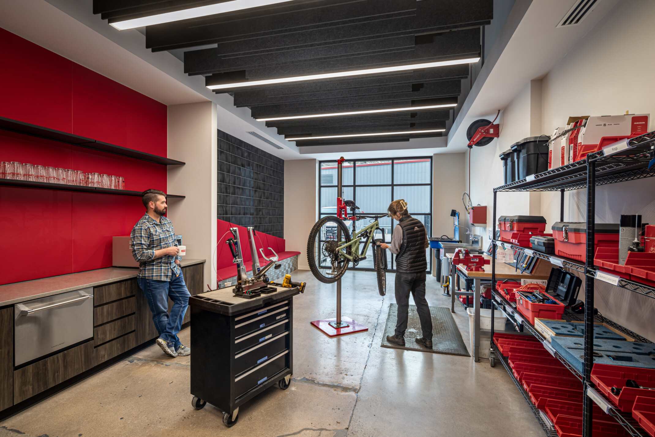 An employee fixes their bike while chatting with a coworker in the bike barn detailed in a bold palette of red and black.