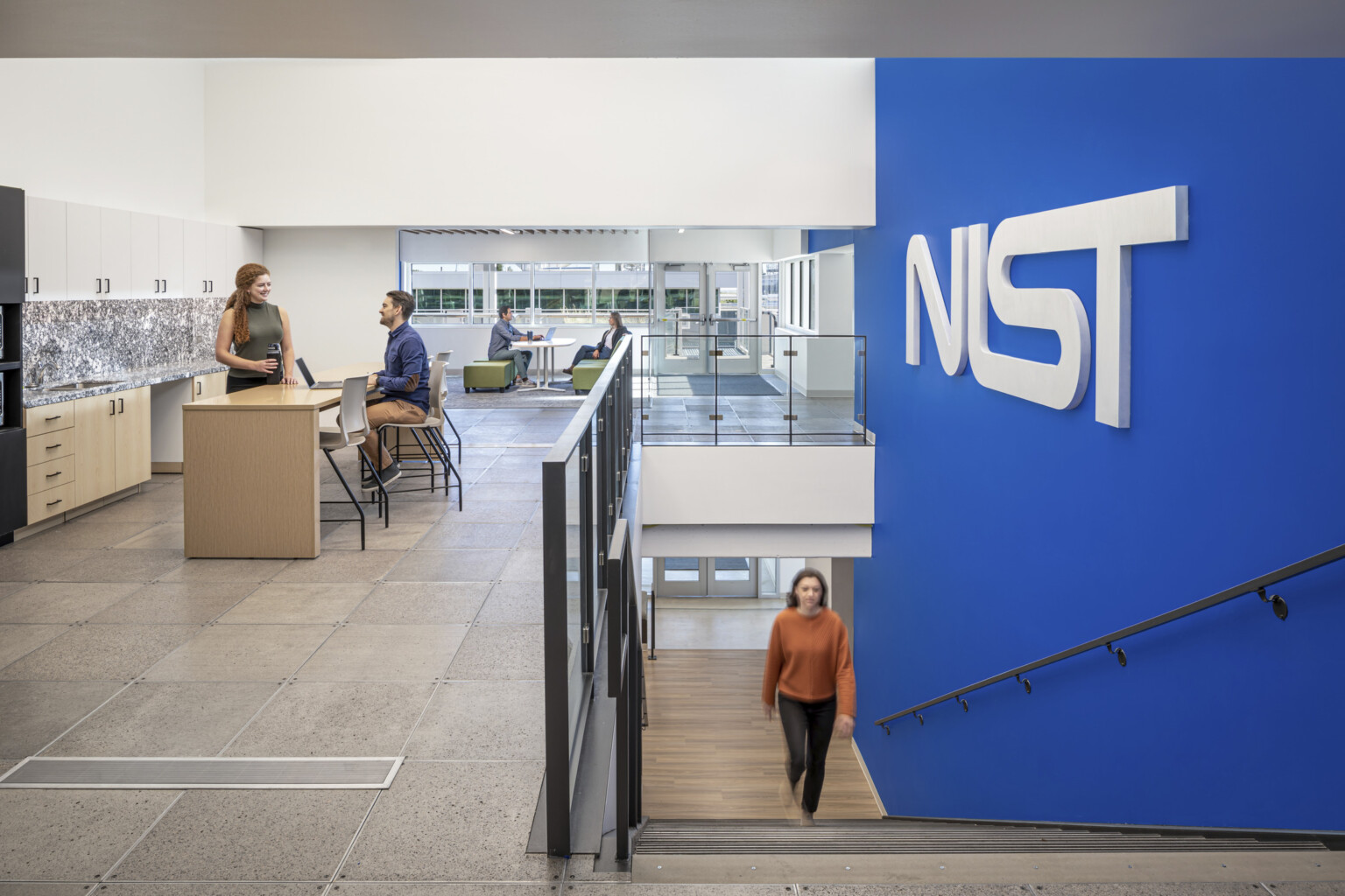 Woman in a orange shirt walking up a staircase with a blue wall to the right with letters NIST. Two couples at the top of the stairs working in collaborative areas