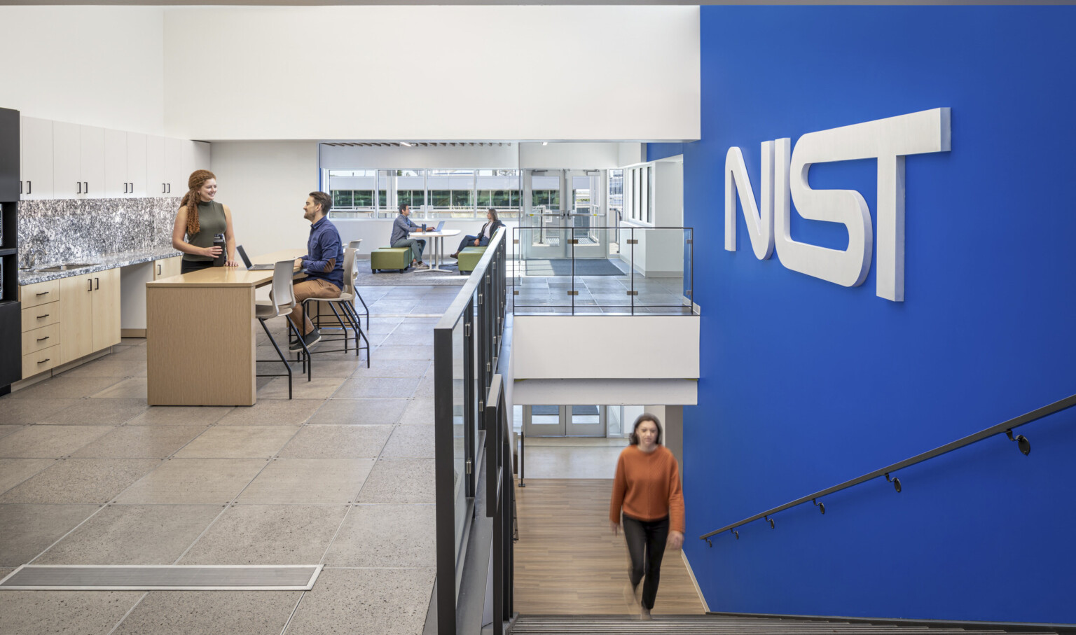Woman in a orange shirt walking up a staircase with a blue wall to the right with letters NIST. Two couples at the top of the stairs working in collaborative areas; NIST Boulder