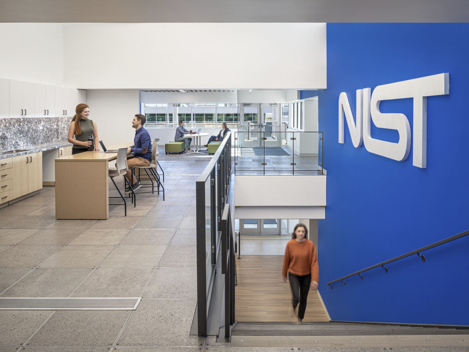 Woman in a orange shirt walking up a staircase with a blue wall to the right with letters NIST. Two couples at the top of the stairs working in collaborative areas