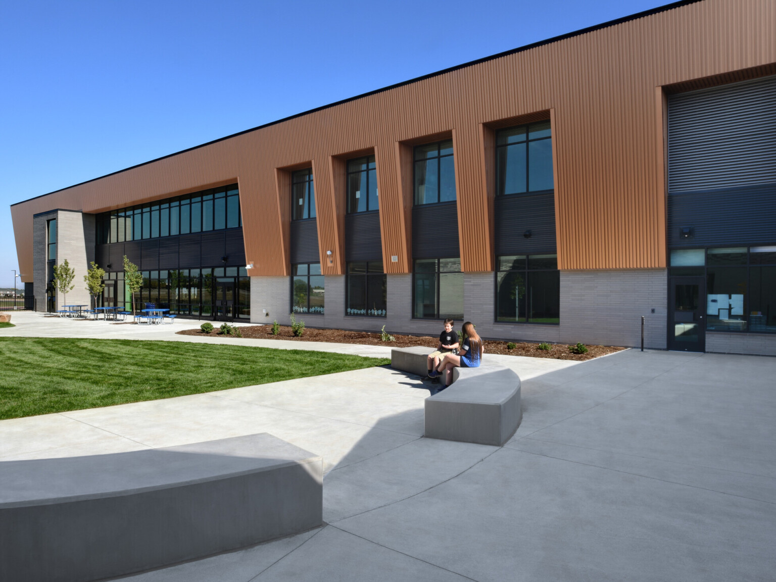 Exterior of two-story engineering academy, brick and wood façade, concrete walkway, grass, outdoor seating, tall windows, under blue skies