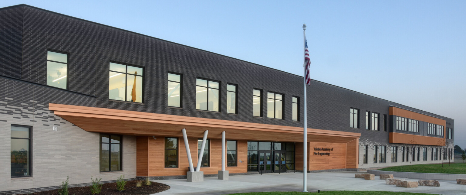 Exterior of two-story engineering academy, walkway, grass, outdoor seating, windows, flag pole with American flag, under blue skies