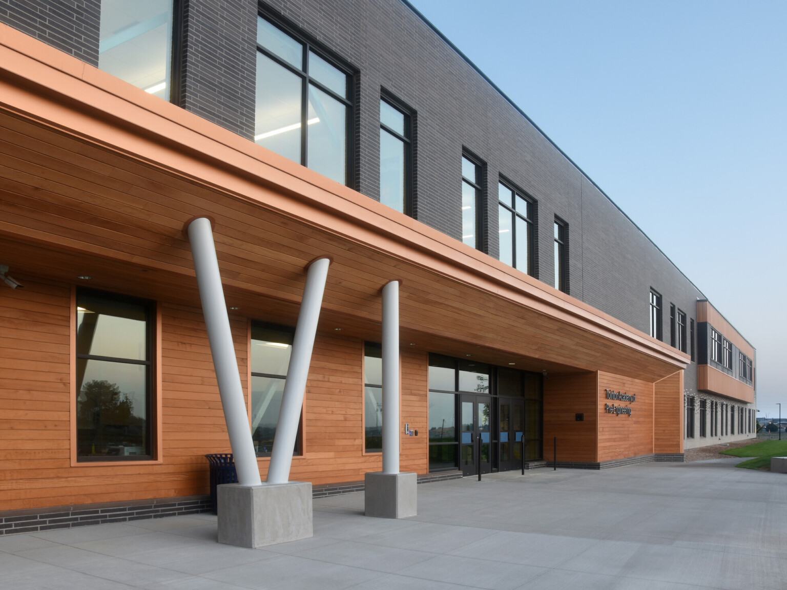 Exterior of two-story engineering academy, brick and wood façade, concrete walkway, grass, outdoor seating, windows, under blue skies