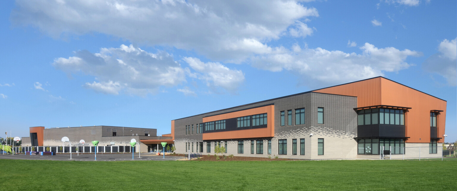 From a distance, building exterior of two-story engineering academy, brick and wood façade, concrete walkway, grass, tall windows, under blue skies and white clouds