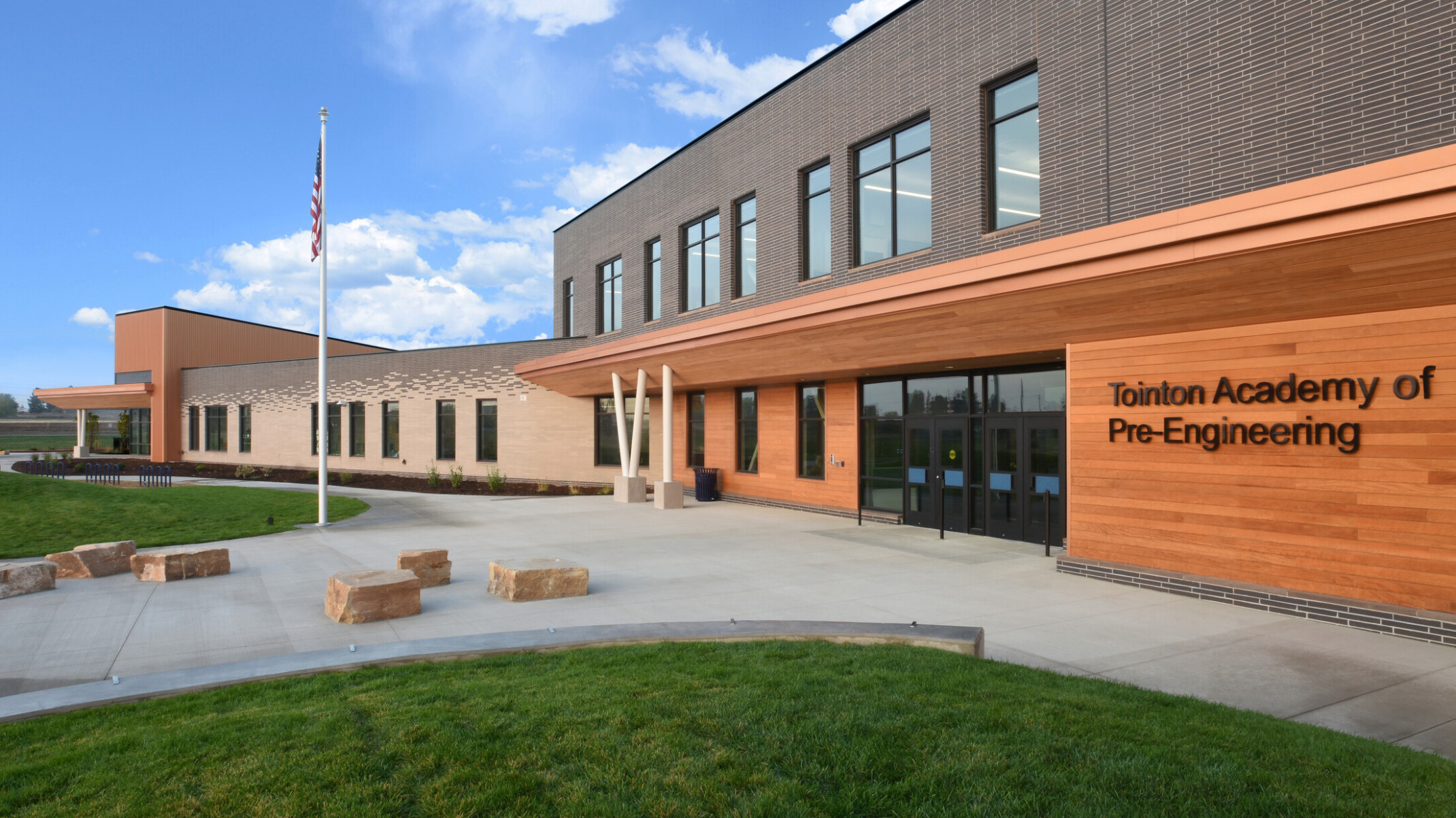 Exterior of two-story engineering academy entrance, walkway, grass, outdoor seating, double-height windows, flag pole with American flag, natural rock seating, under blue skies and fluffy white clouds