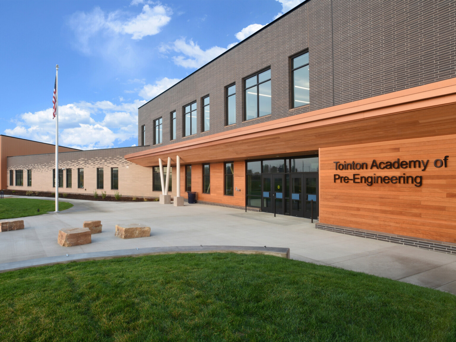 Exterior of two-story engineering academy entrance, walkway, grass, outdoor seating, double-height windows, flag pole with American flag, natural rock seating, under blue skies and fluffy white clouds