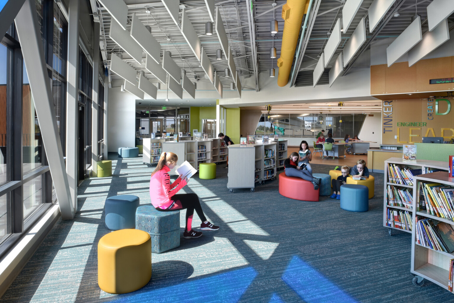 Children in a library with modular furniture and shelves reading books; floor to ceiling windows letting natural light in