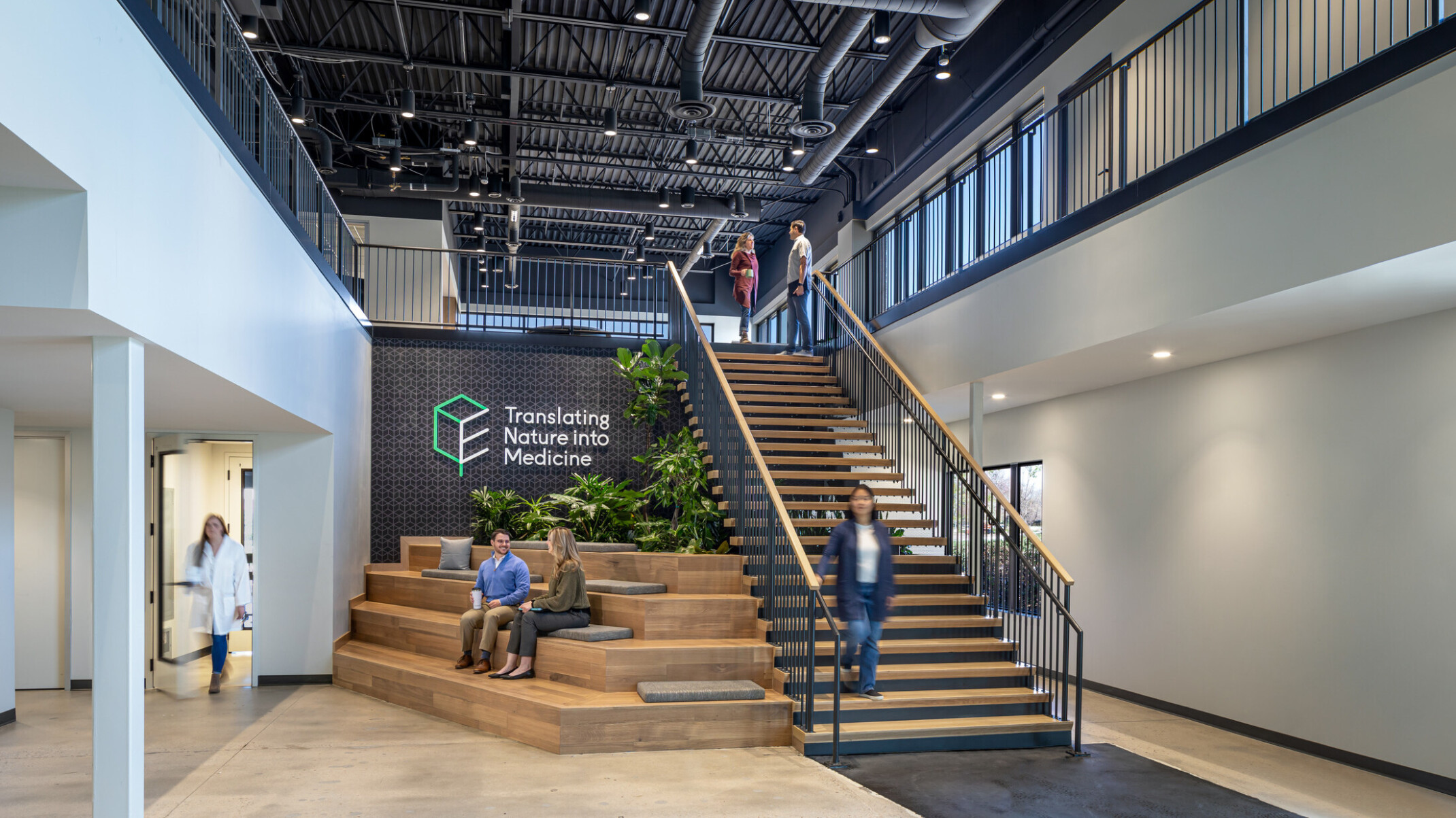 main entrance of a modern laboratory office building; feature staircase with seating built out below and to the left; indoor greenery and exposed structural elements