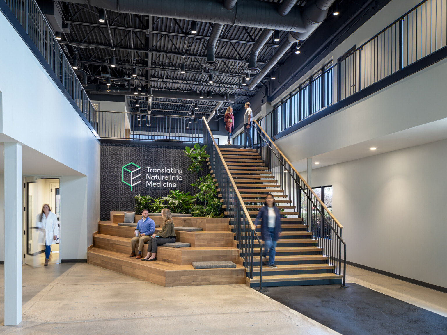 main entrance of a modern laboratory office building; feature staircase with seating built out below and to the left; indoor greenery and exposed structural elements