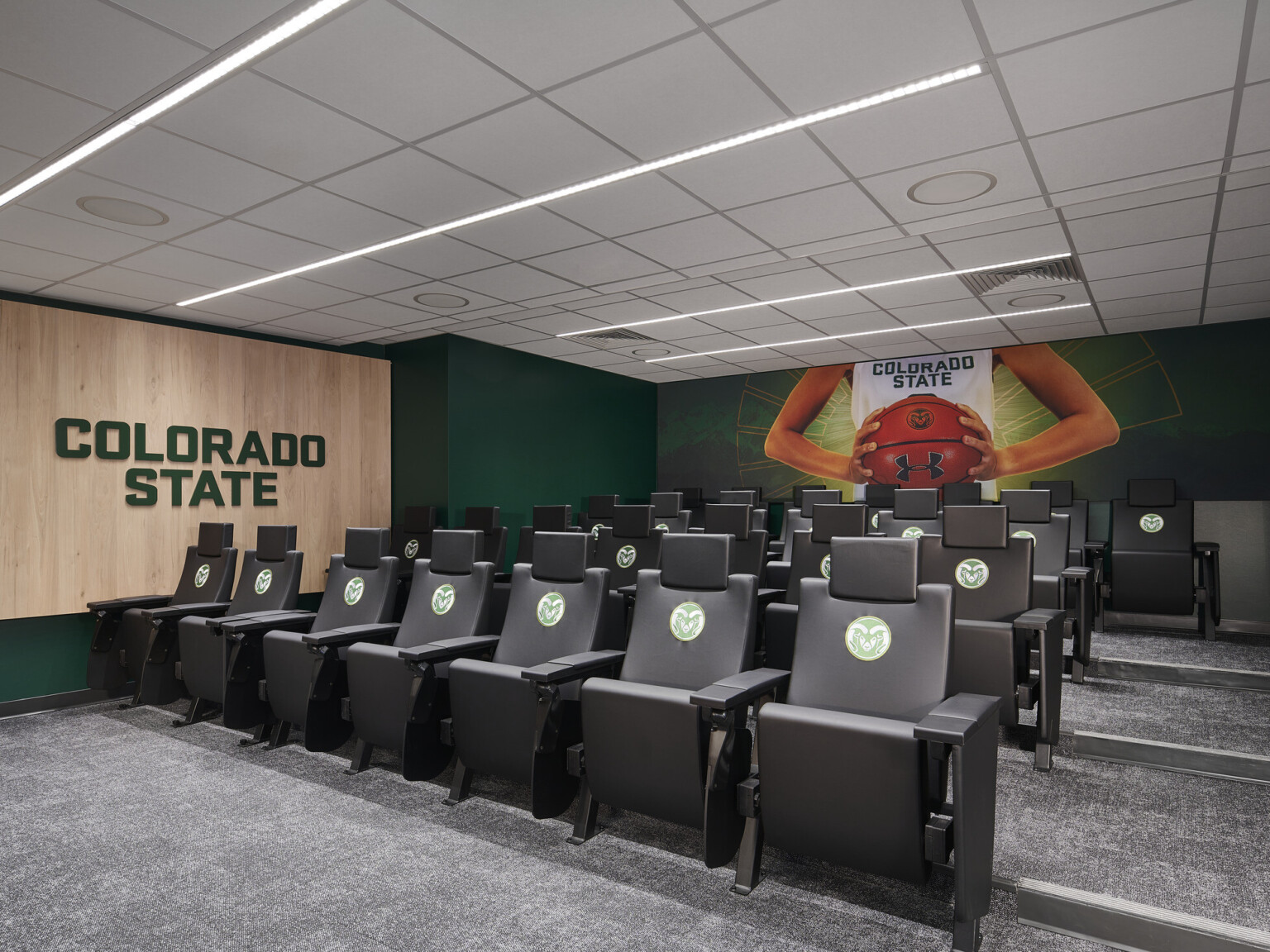 screening room with staggered height branded seating for viewing media, school signage on side wall, large mural of player holding a basketball in team uniform on dark green back wall