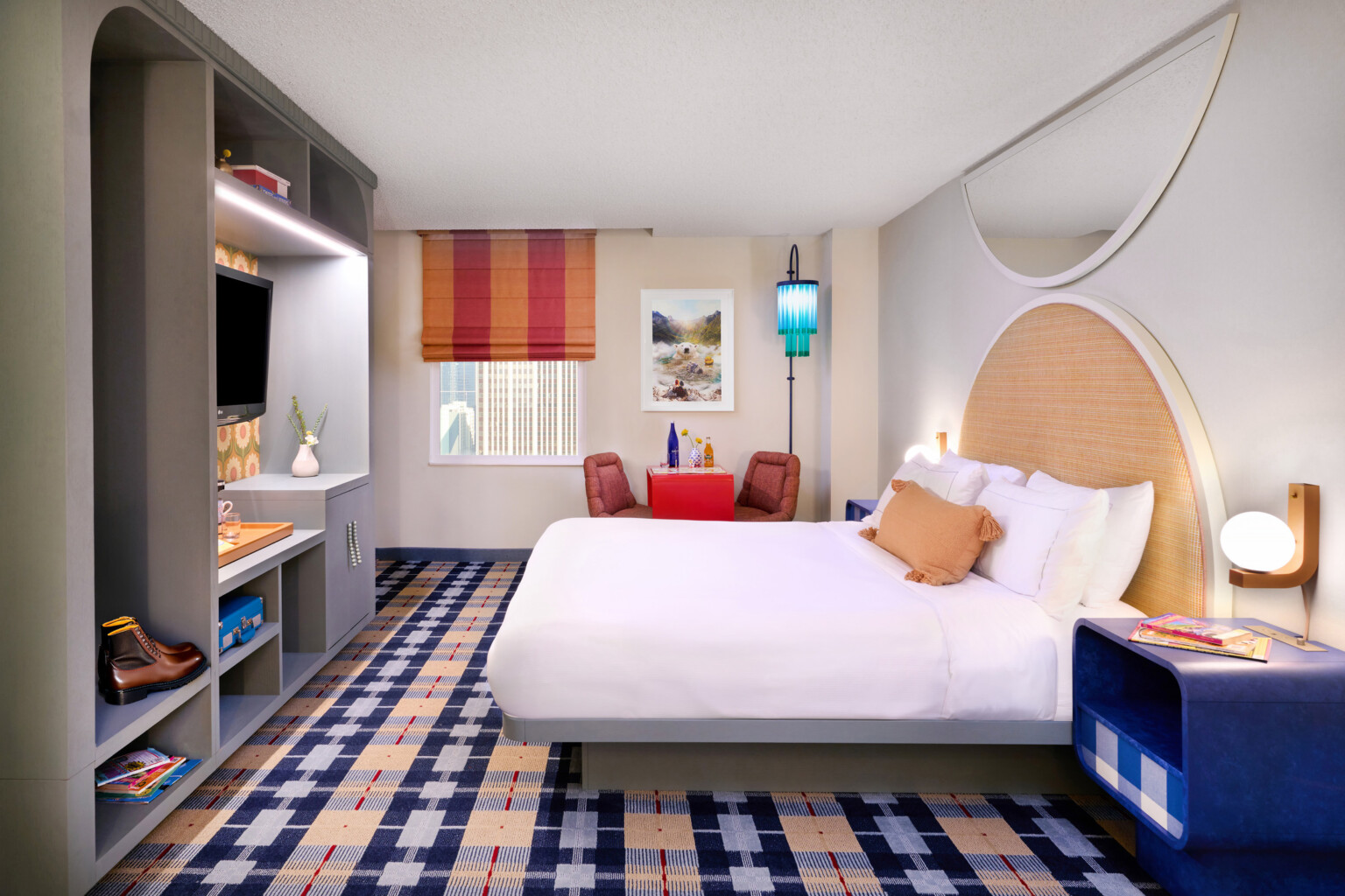 Colorful hotel room; point of view from the entrance door; white bed parallel with the back wall with window and table space; bed footer faces tv, storage space; colorful plaid carpeting