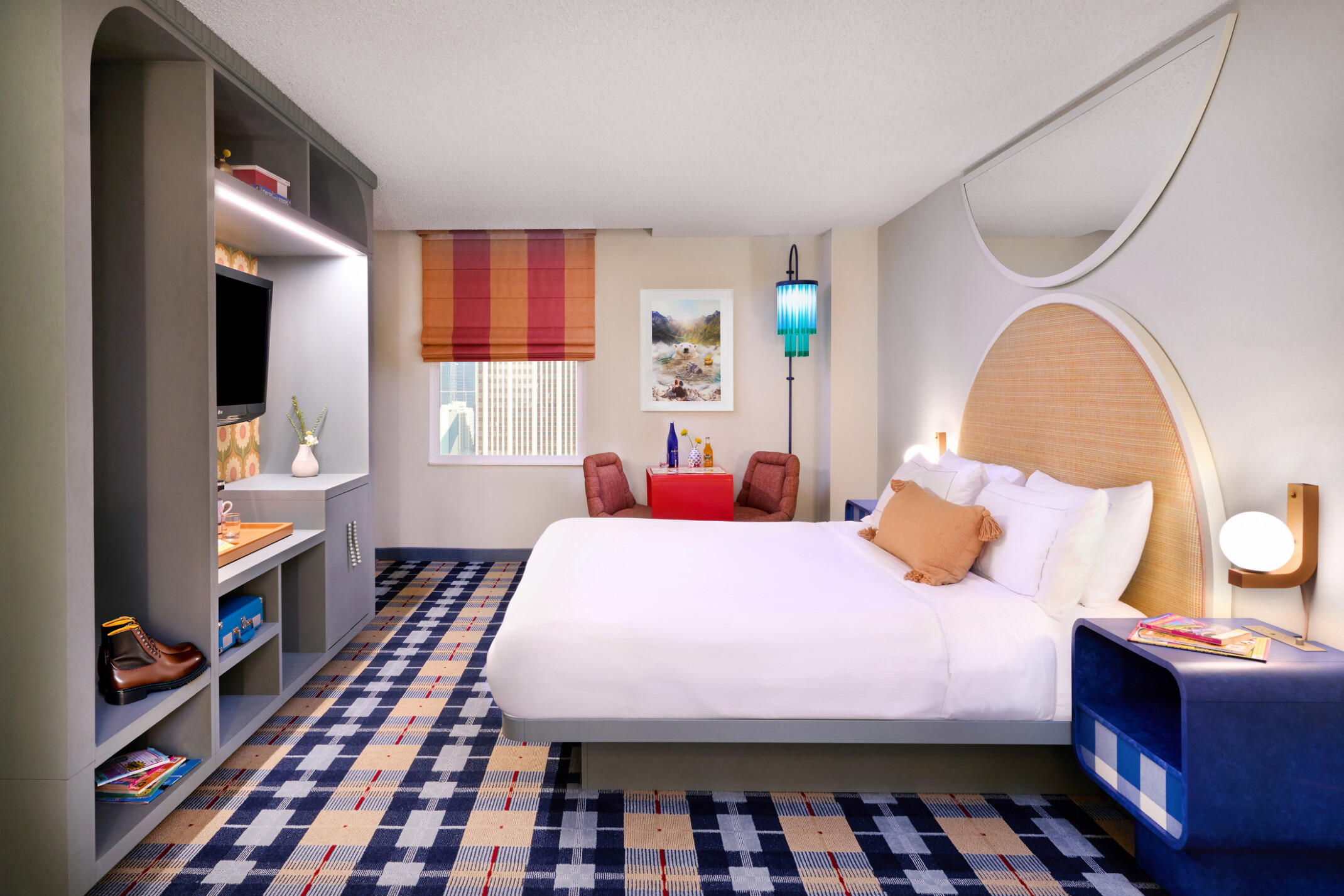 Colorful hotel room; point of view from the entrance door; white bed parallel with the back wall with window and table space; bed footer faces tv, storage space; colorful plaid carpeting