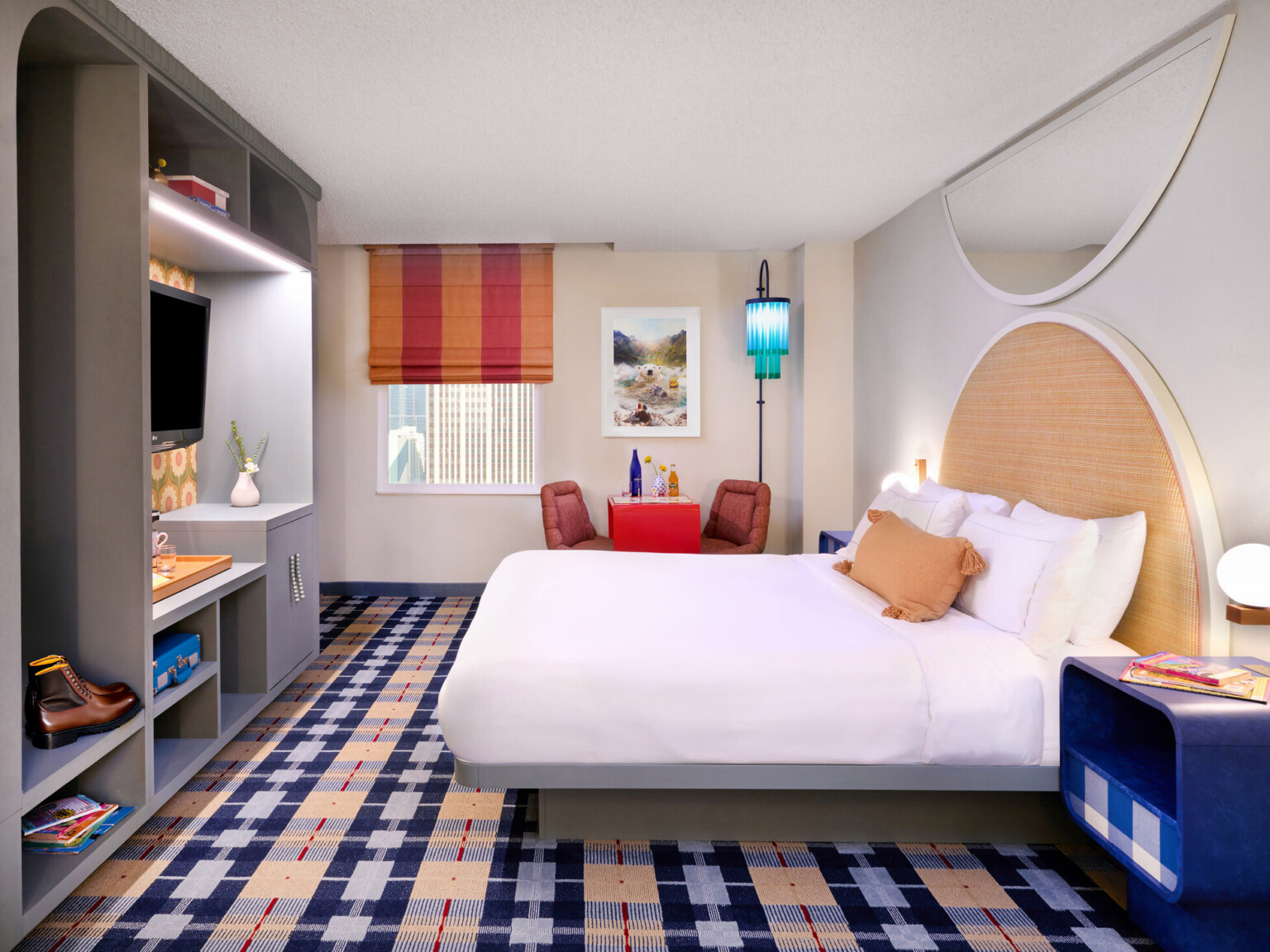 Colorful hotel room; point of view from the entrance door; white bed parallel with the back wall with window and table space; bed footer faces tv, storage space; colorful plaid carpeting