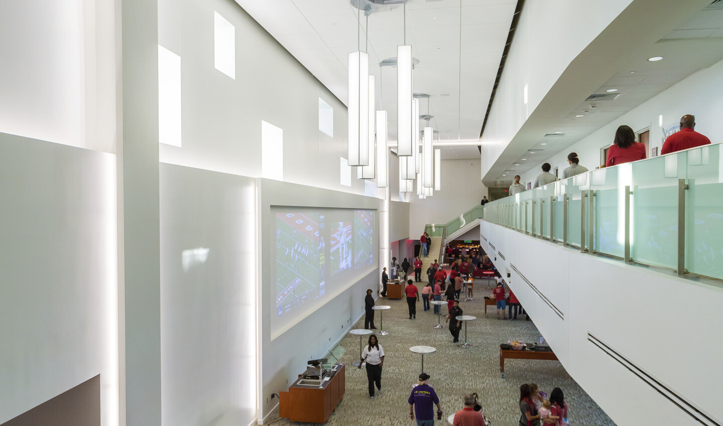 Interior double height room with tables on 1st floor, 2nd floor walkway with glass rail, right. Sculptural chandeliers hang
