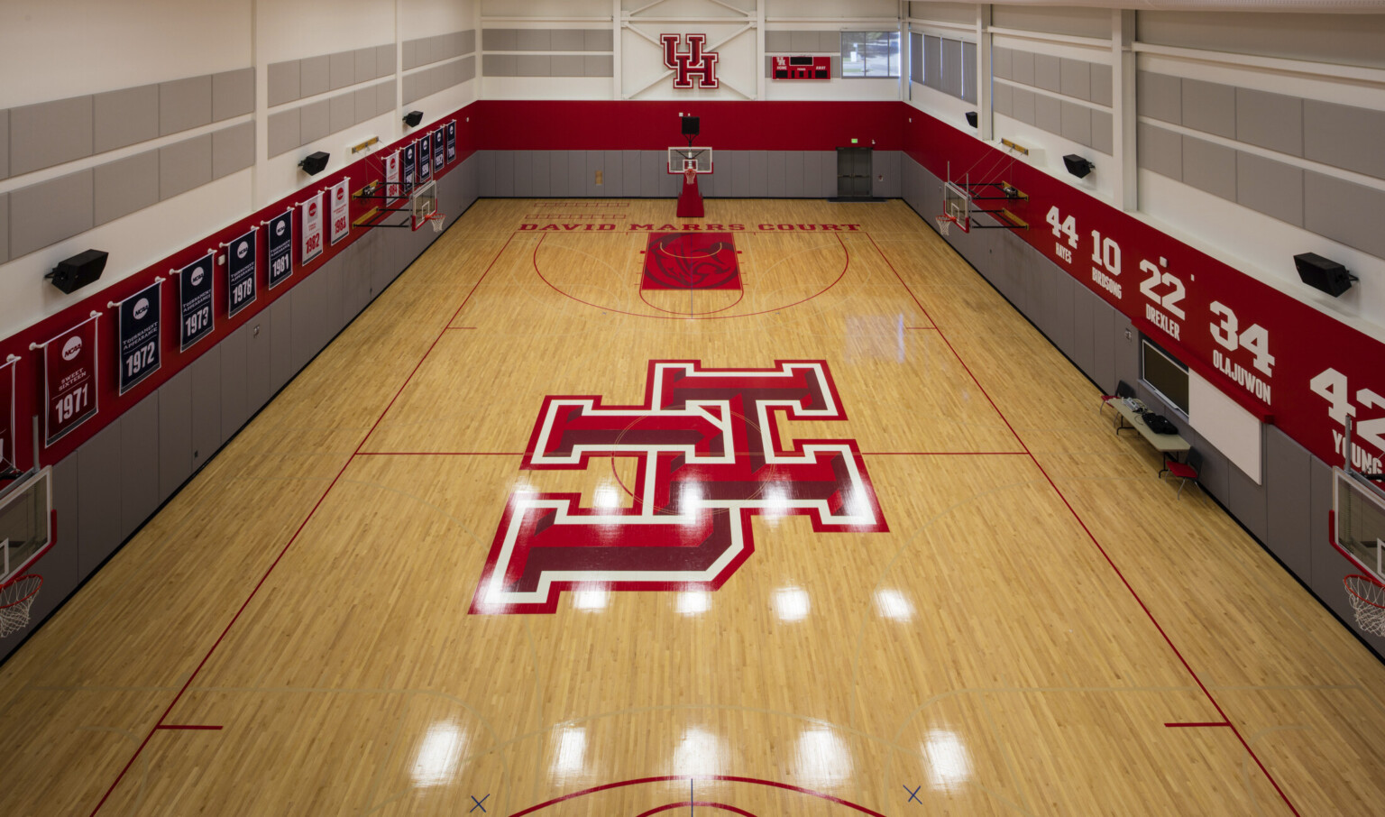 Wood practice court with basketball hoops along sides and UH logo at center. Grey and red stripes on walls with banners left