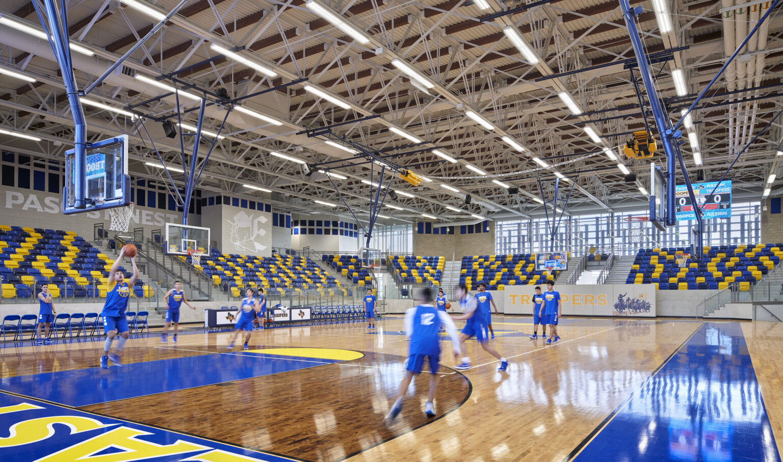 Wood basketball court surrounded by blue and yellow bleachers. Murals with Troopers mascot. Exposed structural beams above