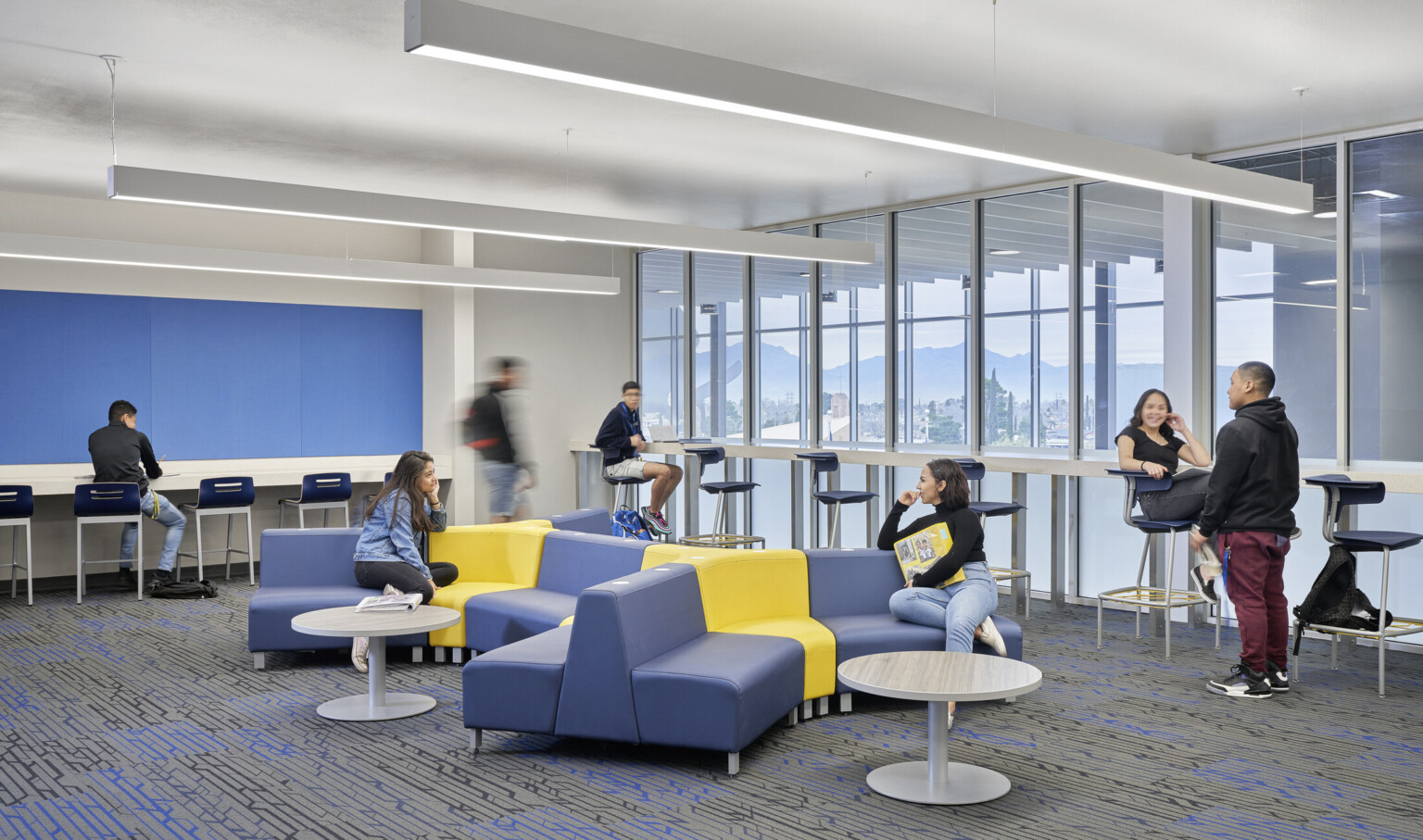 Ergonomic blue and yellow couches in a room with counters and stools along walls. Right wall has floor to ceiling windows