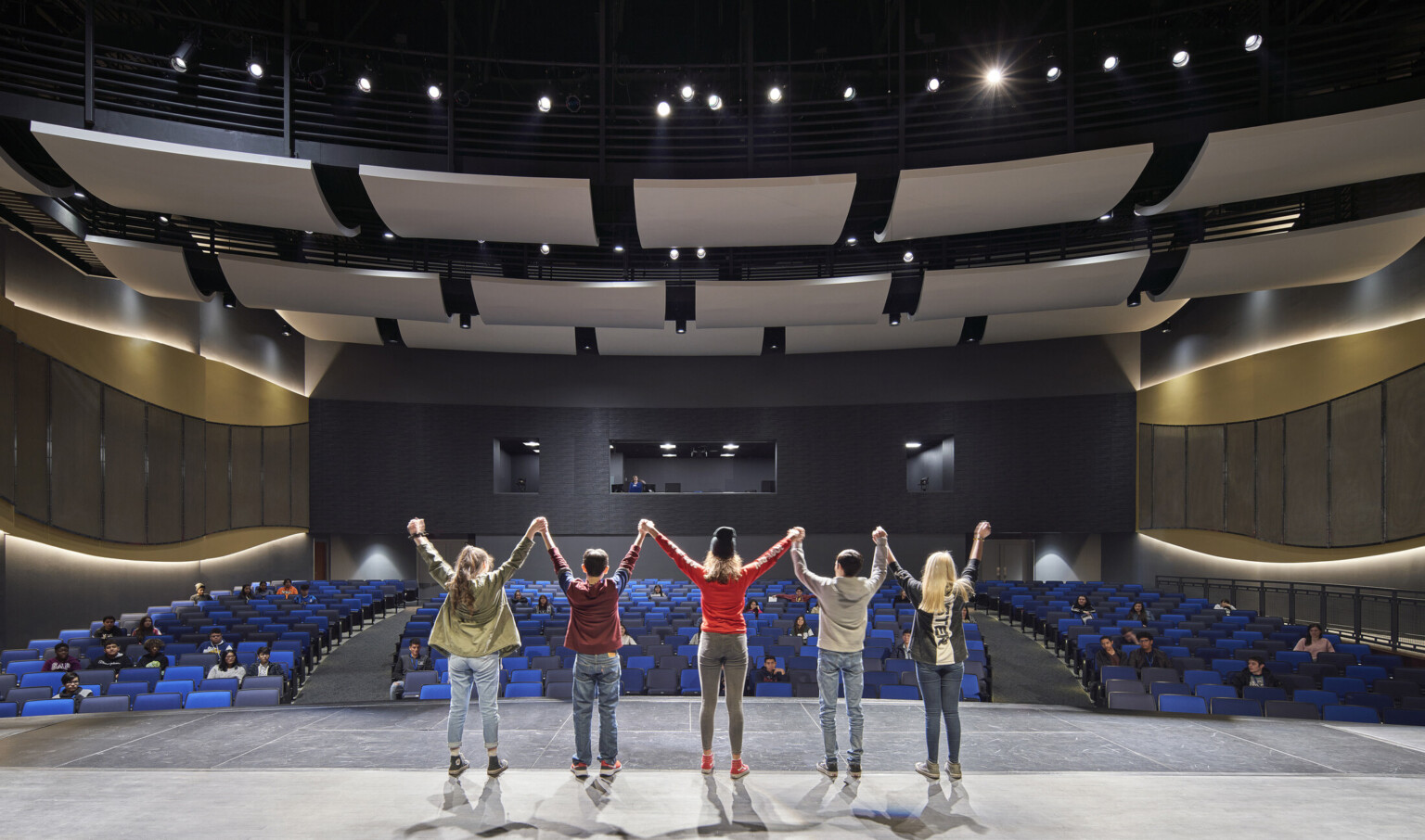 Students stand on stage facing audience. Curved acoustic panels above. Wood details arch along side walls with lights behind