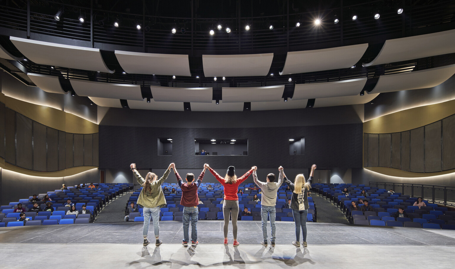Students stand on stage facing audience. Curved acoustic panels above. Wood details arch along side walls with lights behind