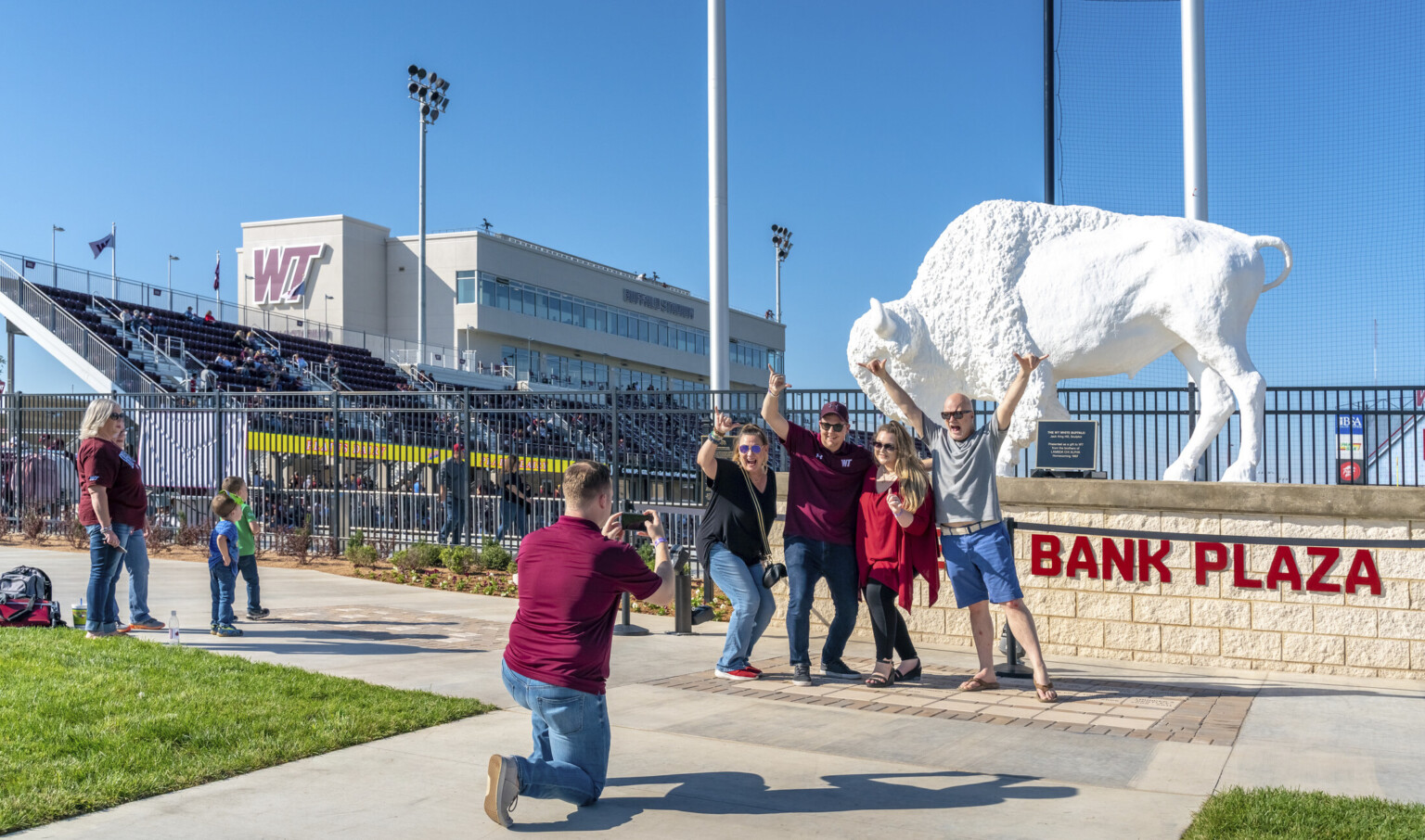 White buffalo sculpture on stone platform outside of black metal stadium fence. Behind, bleachers and box seats are visible