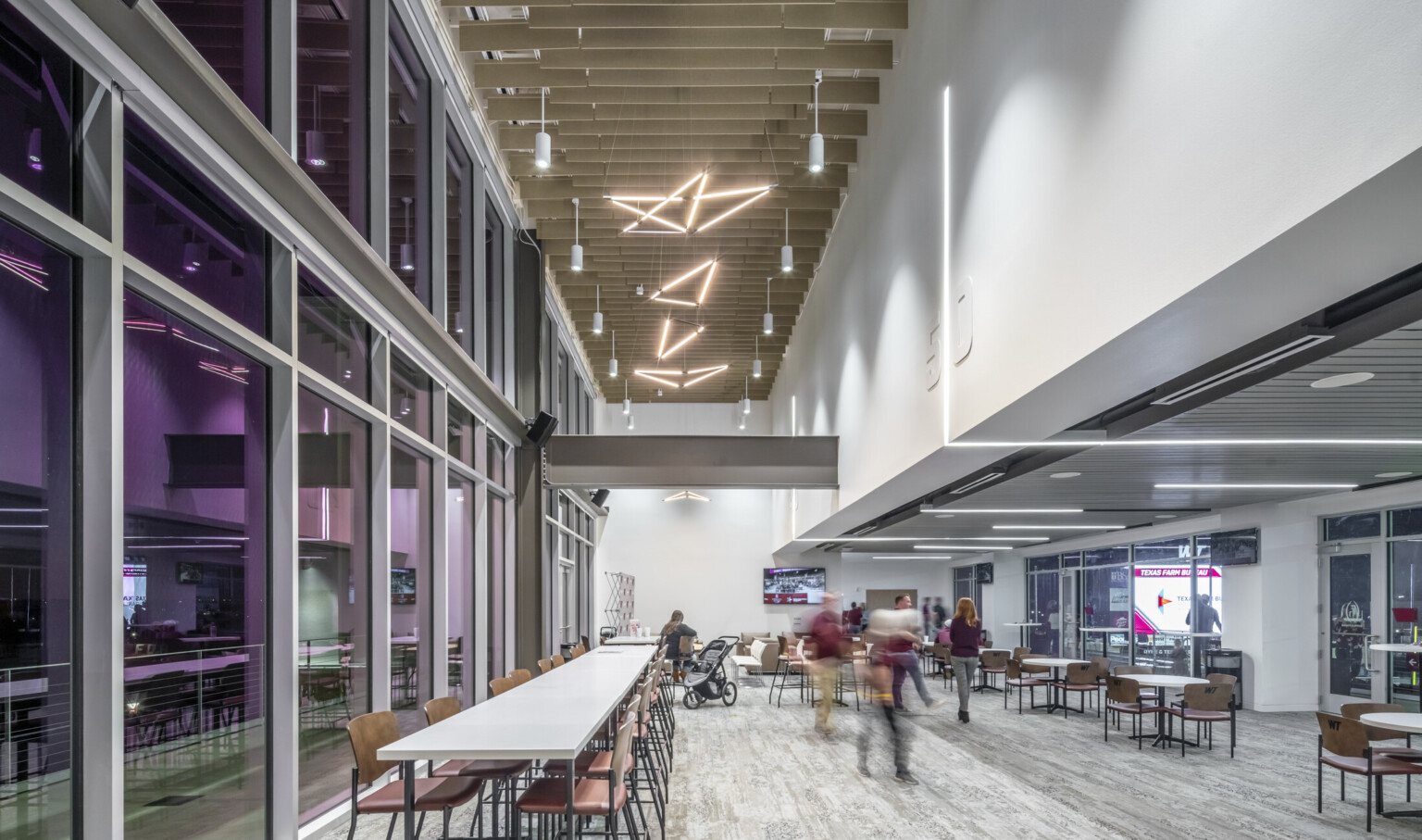 Double height hallway with wood slat ceiling and sculpture light fixtures. Purple translucent windows to left, seating areas