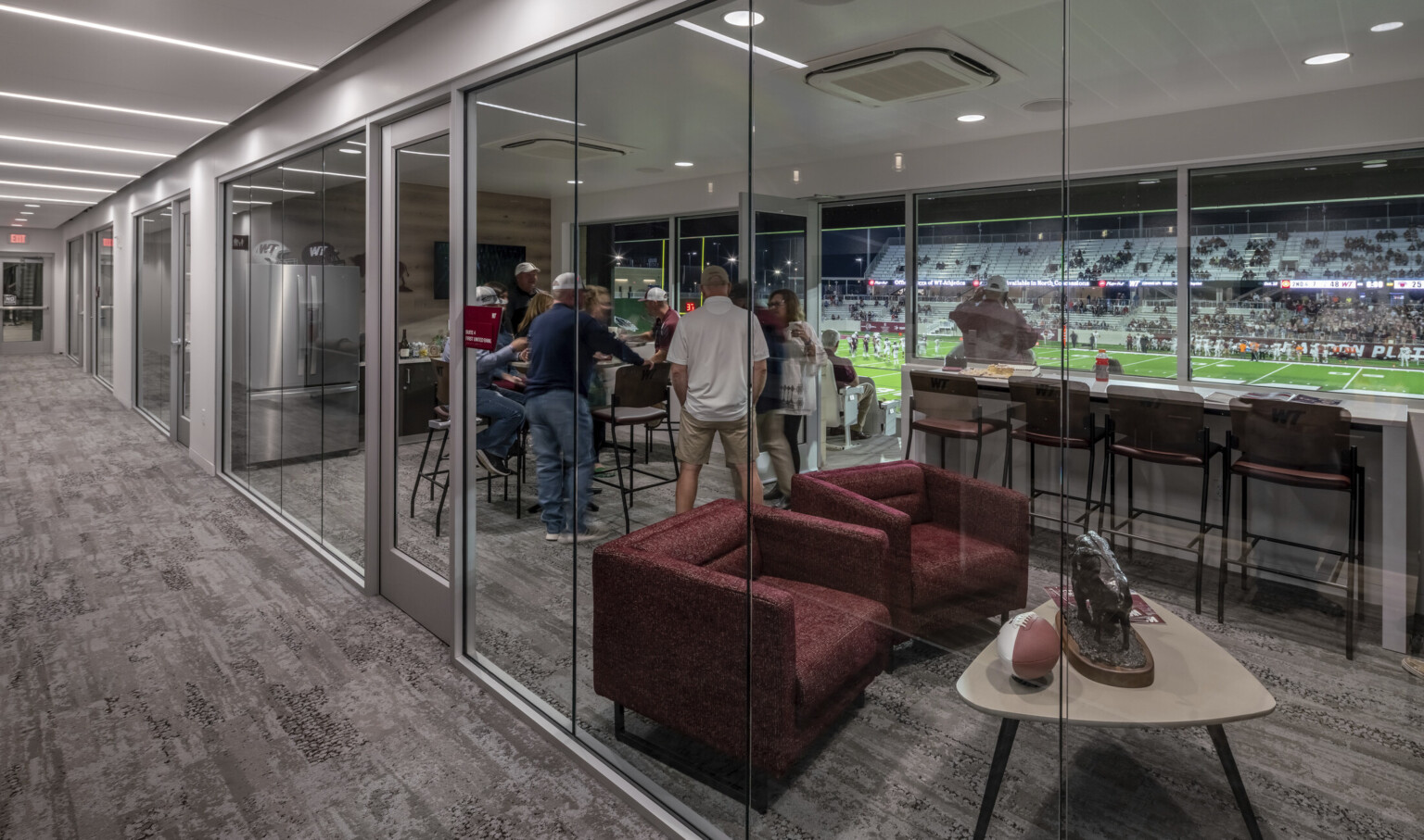 Private suite with glass walls to hallway and field. Inside, armchairs and tables, refrigerator and counter space on far wall