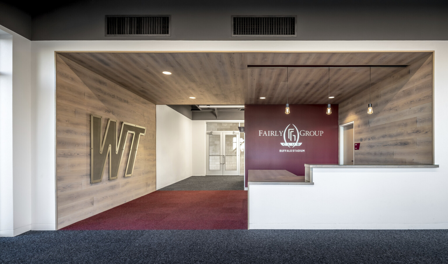 Wood panel room with WT logo on left and red accent wall on right at back with Fairly Group Club logo. Pendant lights above