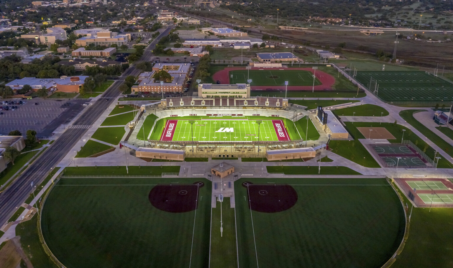 Aerial view of stadium with WT logo in white at field center. Soccer field behind and basketball courts right, buildings left