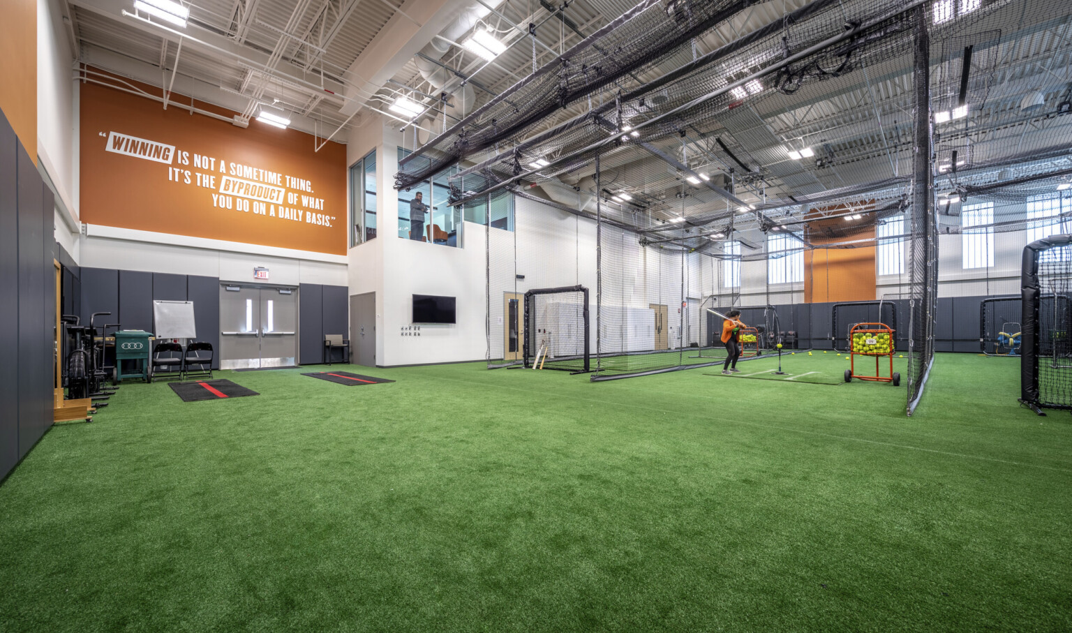 Indoor training space with turf flooring and netted batting cages to the right. Windows to 2nd floor viewing room by mural