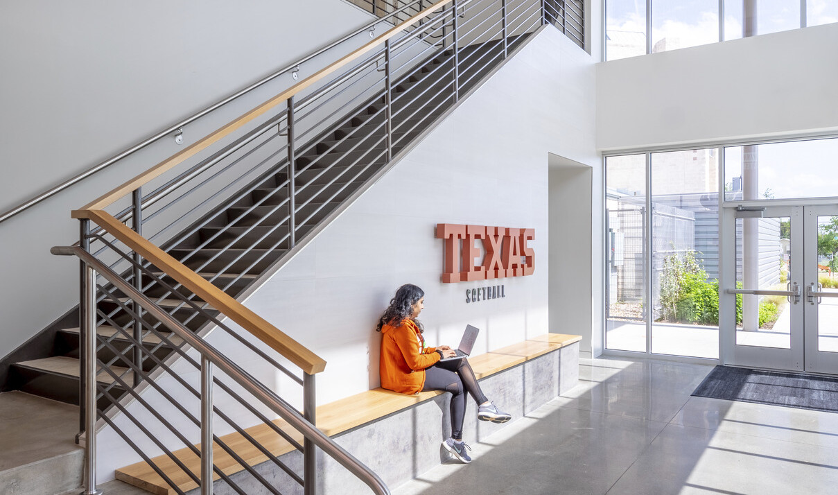 Wood bench along white wall of stair with Texas Softball sign. Glass wall and door leading to exterior walkway