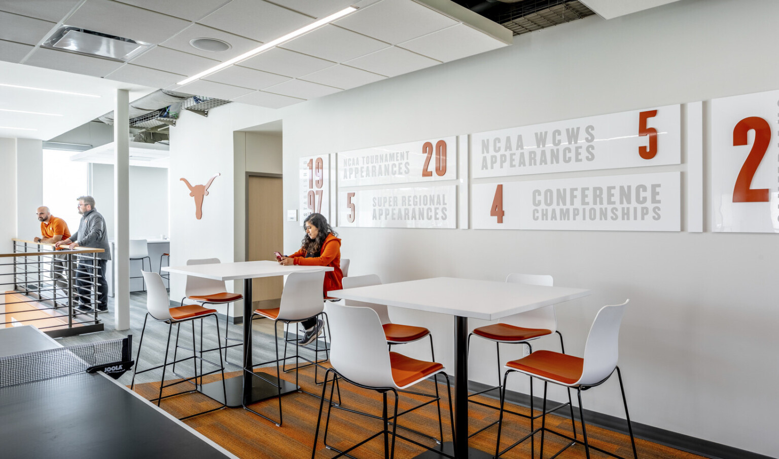 Room off hallway with high top tables and chairs along grey wall with white signs of team facts. Ping pong table to left.
