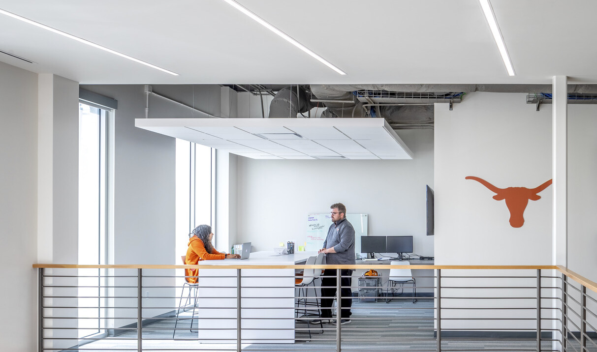 Tall white room with hanging panel at the end of hallway. Counters and chairs at center, computers and white board along wall