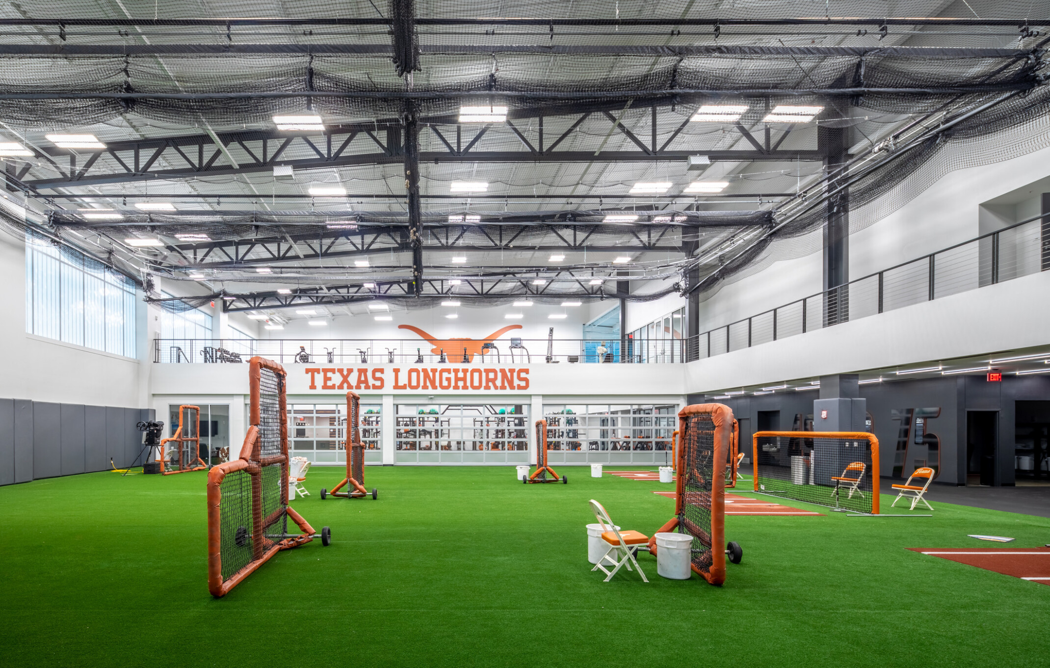 Indoor baseball and softball training facility with artificial turf, multiple batting and pitching stations, overhead netting, and a mezzanine level overlooking the practice area. A large "TEXAS LONGHORNS" sign and Longhorn logo are displayed on the far wall