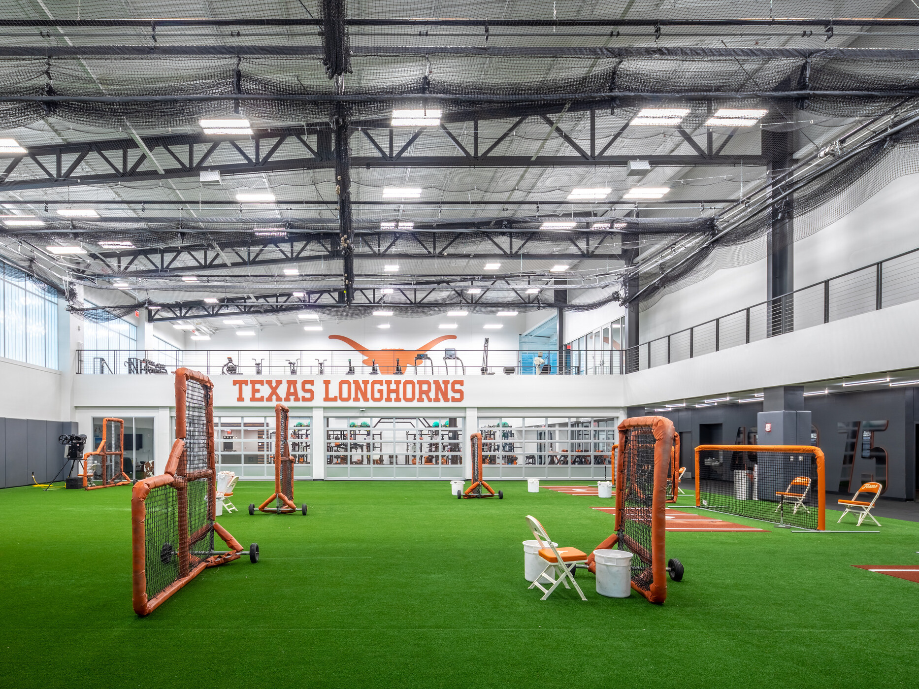 Indoor baseball and softball training facility with artificial turf, multiple batting and pitching stations, overhead netting, and a mezzanine level overlooking the practice area. A large 