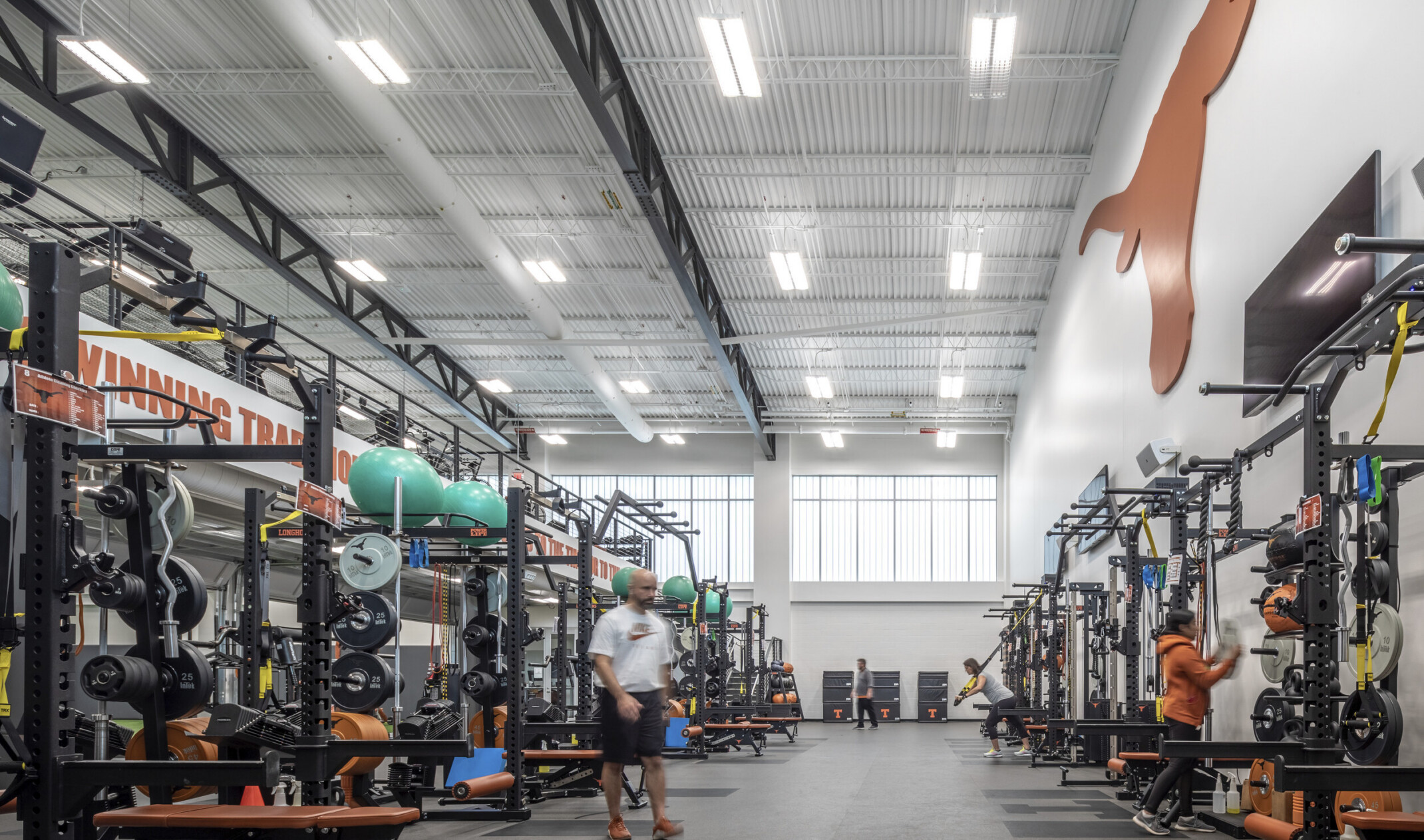 Training room with weight equipment along both walls. Double height white room with more equipment on 2nd floor balcony
