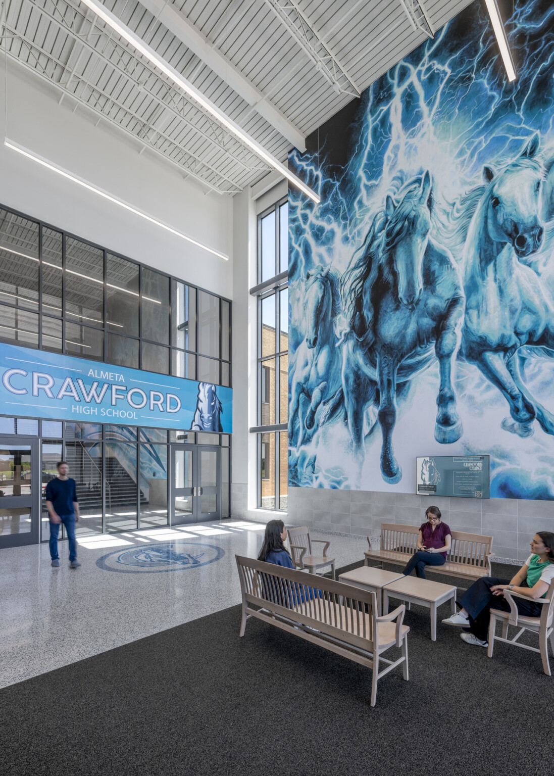 Seating area with large mural in shades of blue behind benches. Large wall filled with windows with artwork with words Almeta Crawford High school in the middle Seating area with large mural in shades of blue behind benches. Large wall filled with windows with artwork with words Almeta Crawford High school in the middle