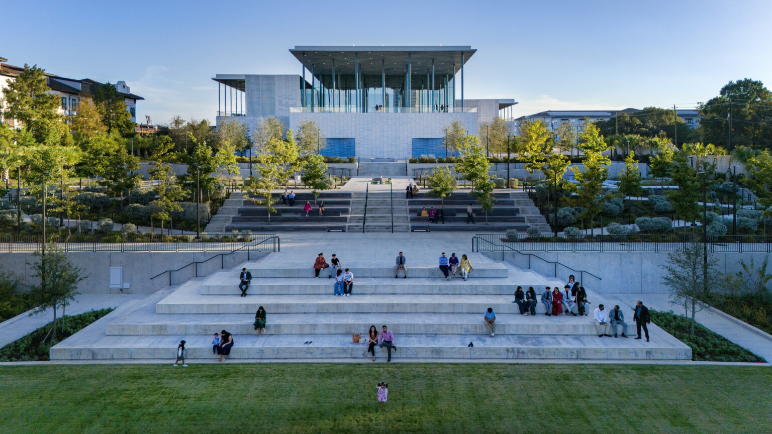 A modern building with a glass façade sits atop stone steps surrounded by lush greenery.