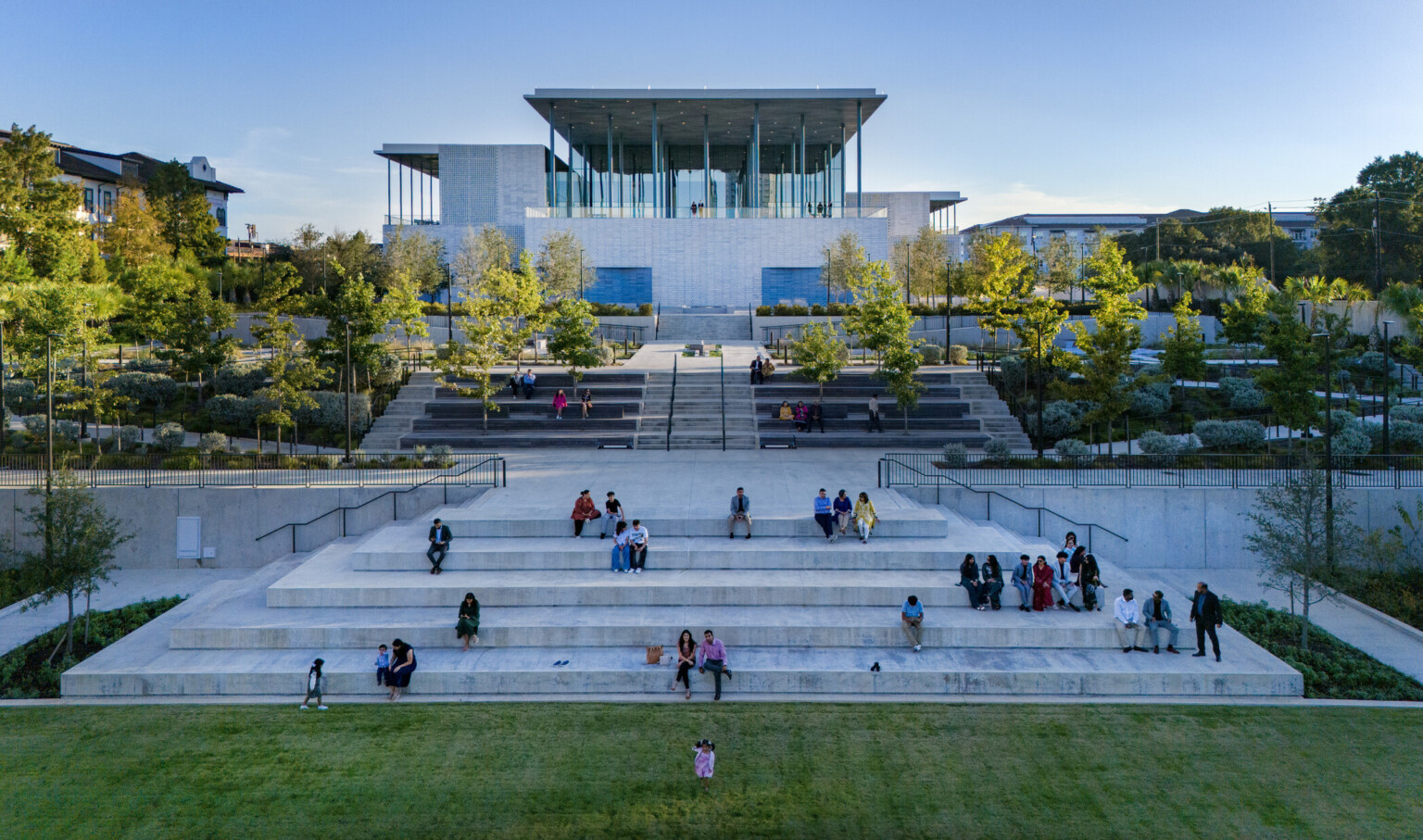 A modern building with a glass façade sits atop stone steps surrounded by lush greenery.