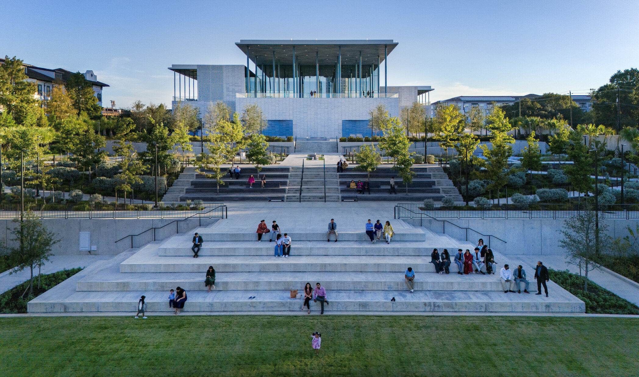 A modern building with a glass façade sits atop stone steps surrounded by lush greenery.