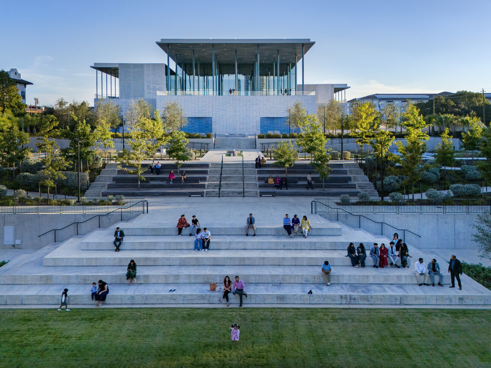 A modern building with a glass façade sits atop stone steps surrounded by lush greenery.