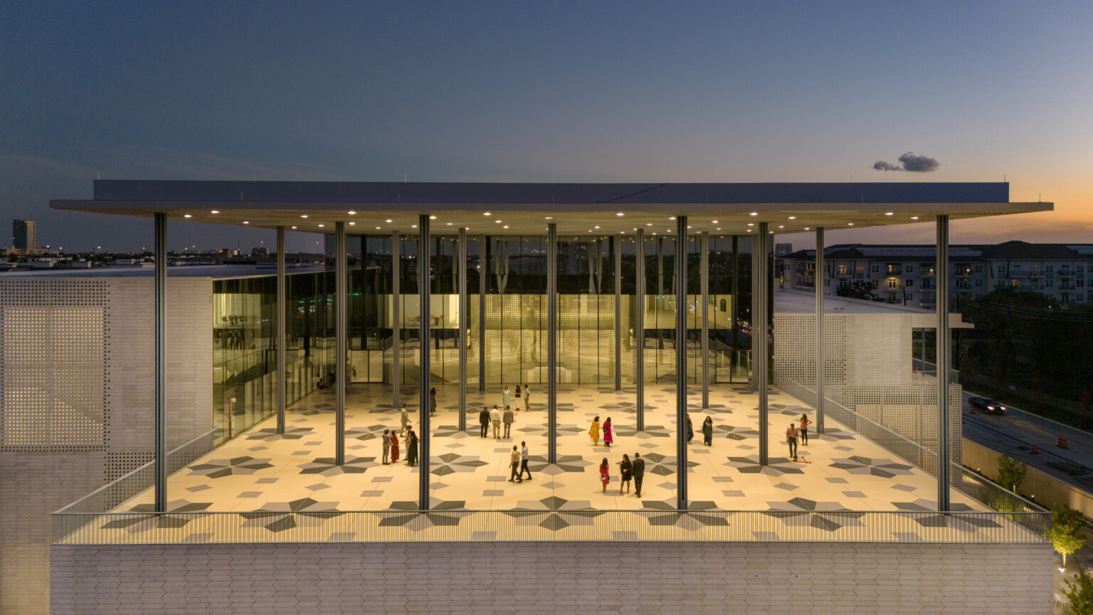 A modern building at sunset with a large, covered veranda supported by columns. Geometric patterns adorn tile floors.
