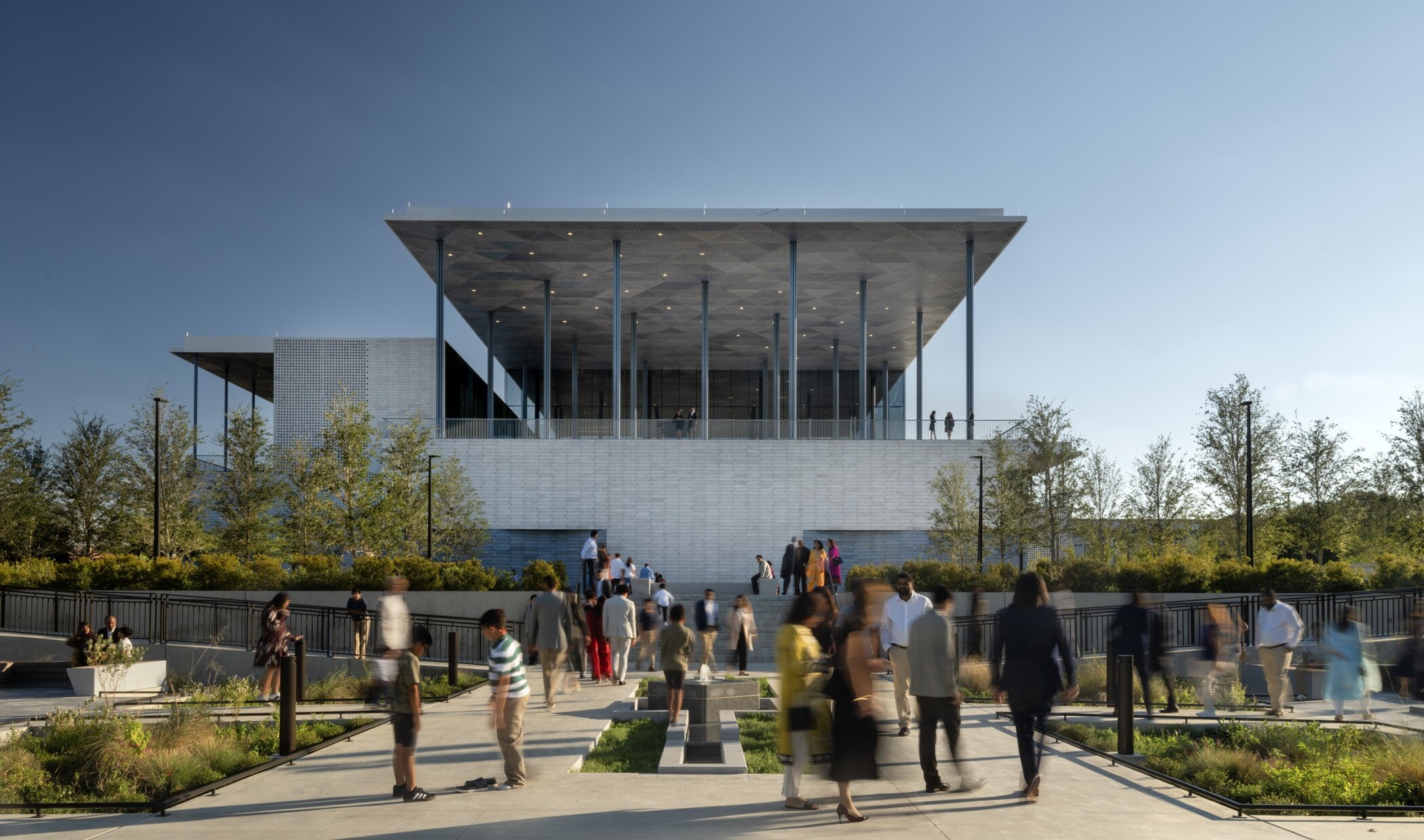 Contemporary building surrounded by landscaped gardens with a fountain in the foreground. Groups of people congregate in the gardens and on an elevated veranda overlooking the gardens.