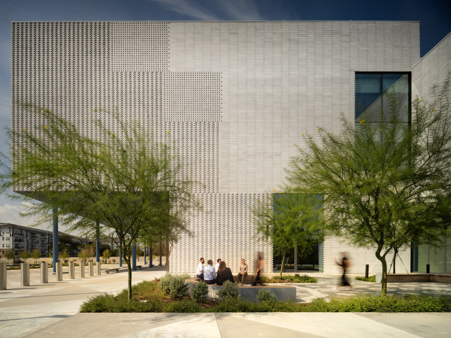 A building exterior clad in light gray/sand colored marble arranged in varying patterns and shapes to evoke a tapestry in stone. An exterior courtyard features flowering trees and plantings, geometric patterned floor tiles, and seating