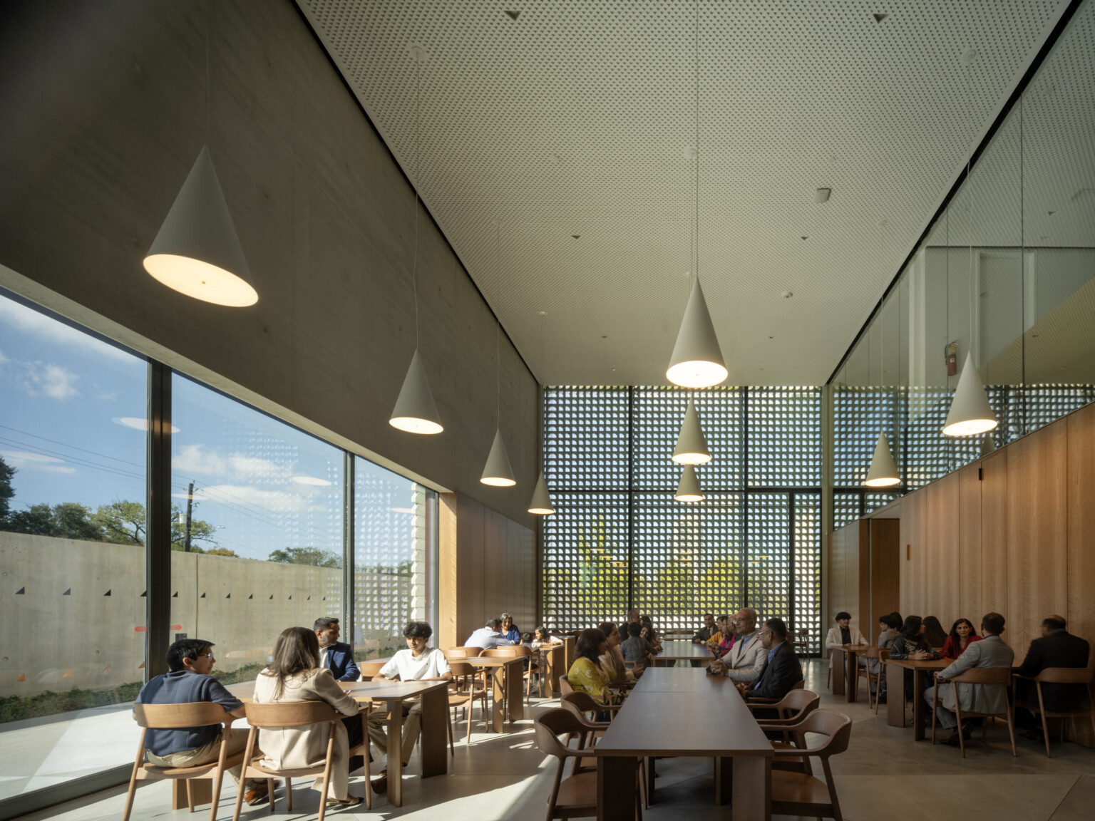 Museum café interior with visitors seated at wooden tables beneath pendant lights. A tall marble screen wall filters natural light on the right, while warm wood paneling lines the back wall.