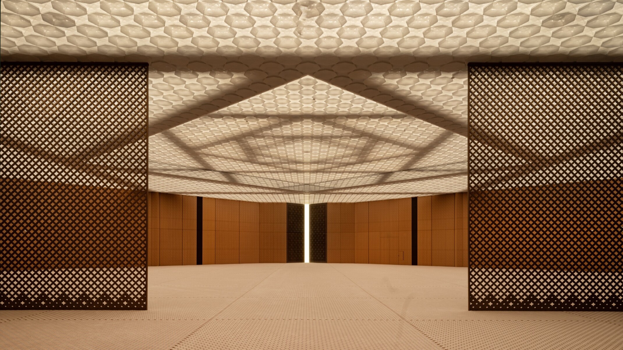 A prayer hall with wooden walls and a ceiling with integrated lighting. The ceiling features layers of patterned, perforated aluminum creating a soft ethereal effect. The structural system of steel beams in interlocking squares is visible above the ceiling.